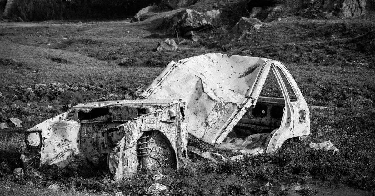 Image en noir et blanc d'une carcasse de voiture rouillée et abandonnée dans un champ herbeux.