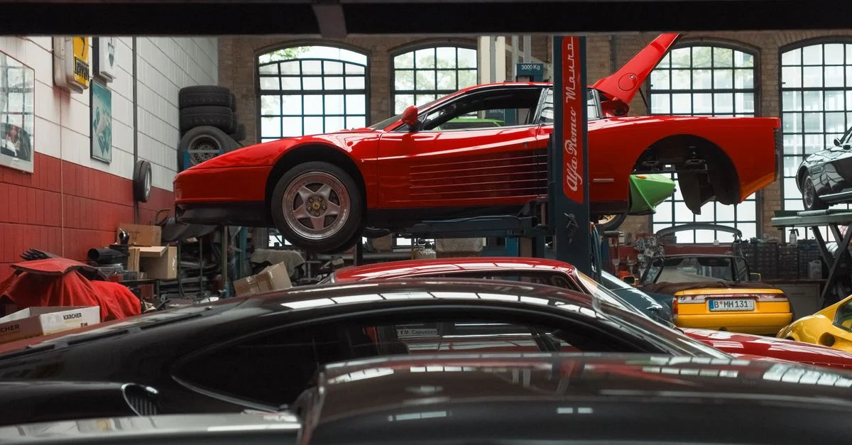 Une voiture de sport rouge vif est surélevée sur un pont élévateur à l'intérieur d'un garage de réparation automobile, entourée de divers véhicules et équipements.