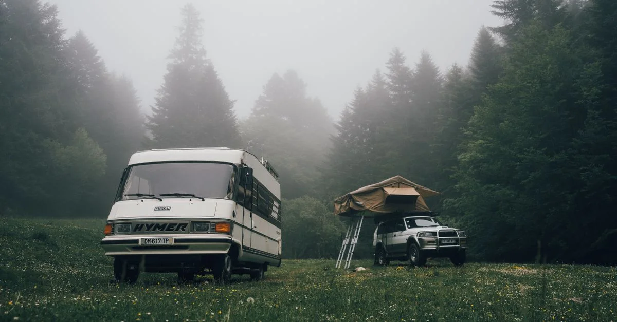 Vue scénique d'un camping-car et d'une tente dans une prairie forestière brumeuse, parfait pour les campeurs aventuriers.