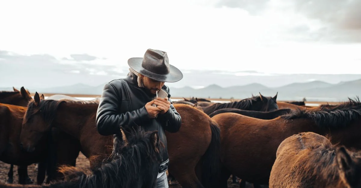Un cowboy en veste en cuir et chapeau allume une cigarette parmi un troupeau de chevaux dans un cadre extérieur pittoresque.
