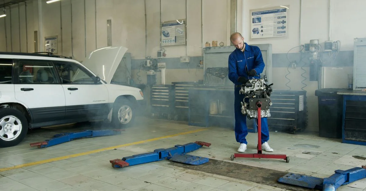 Mécanicien en salopette bleue travaillant sur un moteur de voiture dans un atelier de réparation auto avec une voiture blanche.