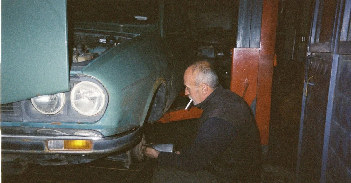 Un mécanicien âgé répare une voiture vintage à l'intérieur, fumant une cigarette tout en travaillant.