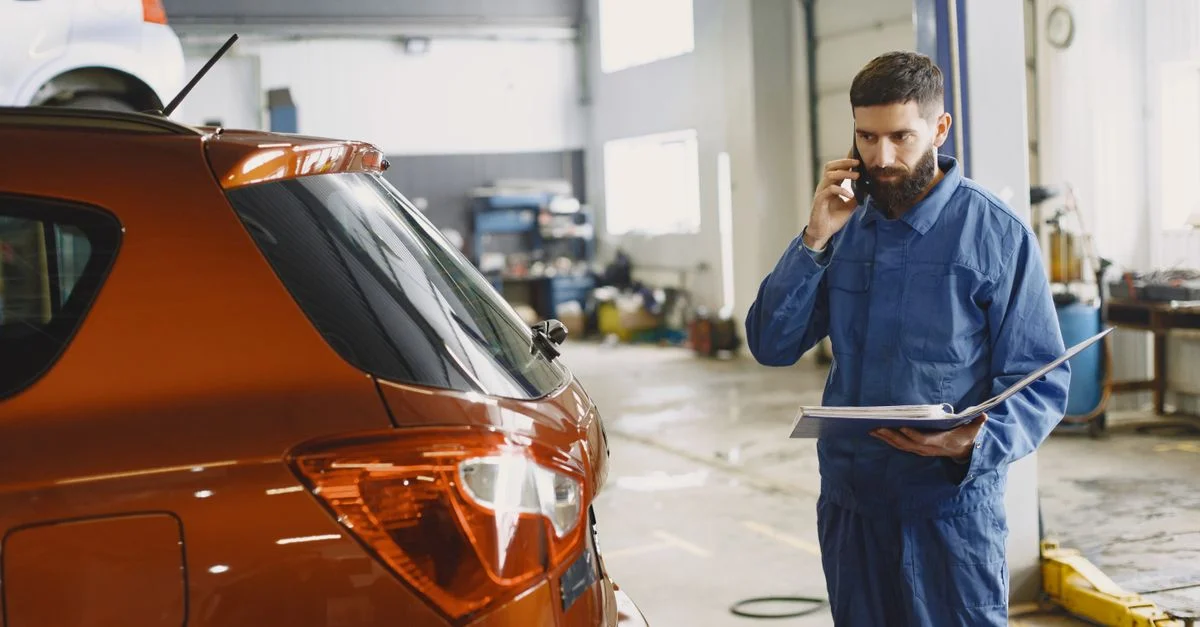Un mécanicien en uniforme bleu parle au téléphone à côté d'une voiture rouge dans un atelier de réparation automobile.