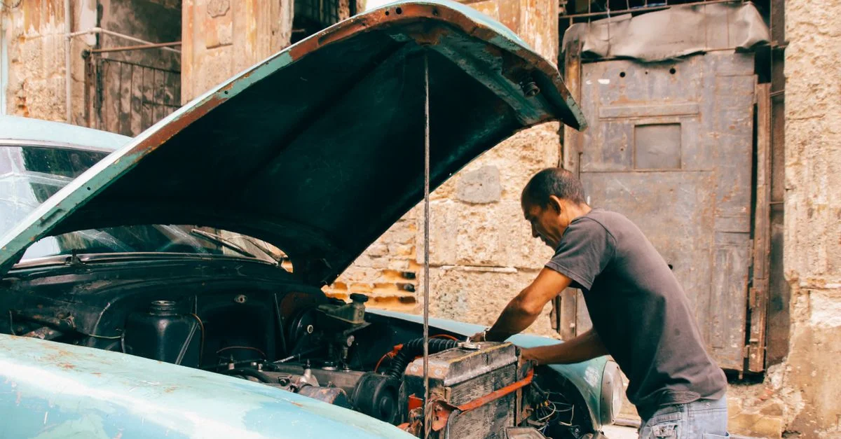Un homme travaillant sur le moteur d'une vieille voiture vintage dans un garage extérieur rustique.