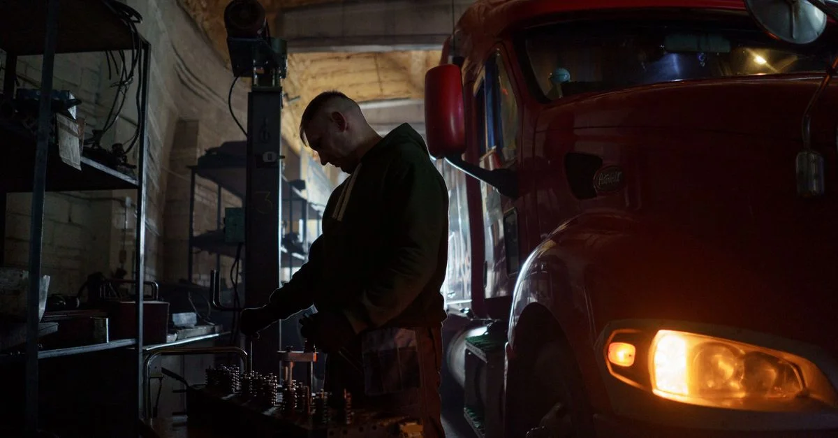 Un mécanicien se concentre sur son travail à côté d'un grand camion rouge dans un atelier faiblement éclairé.