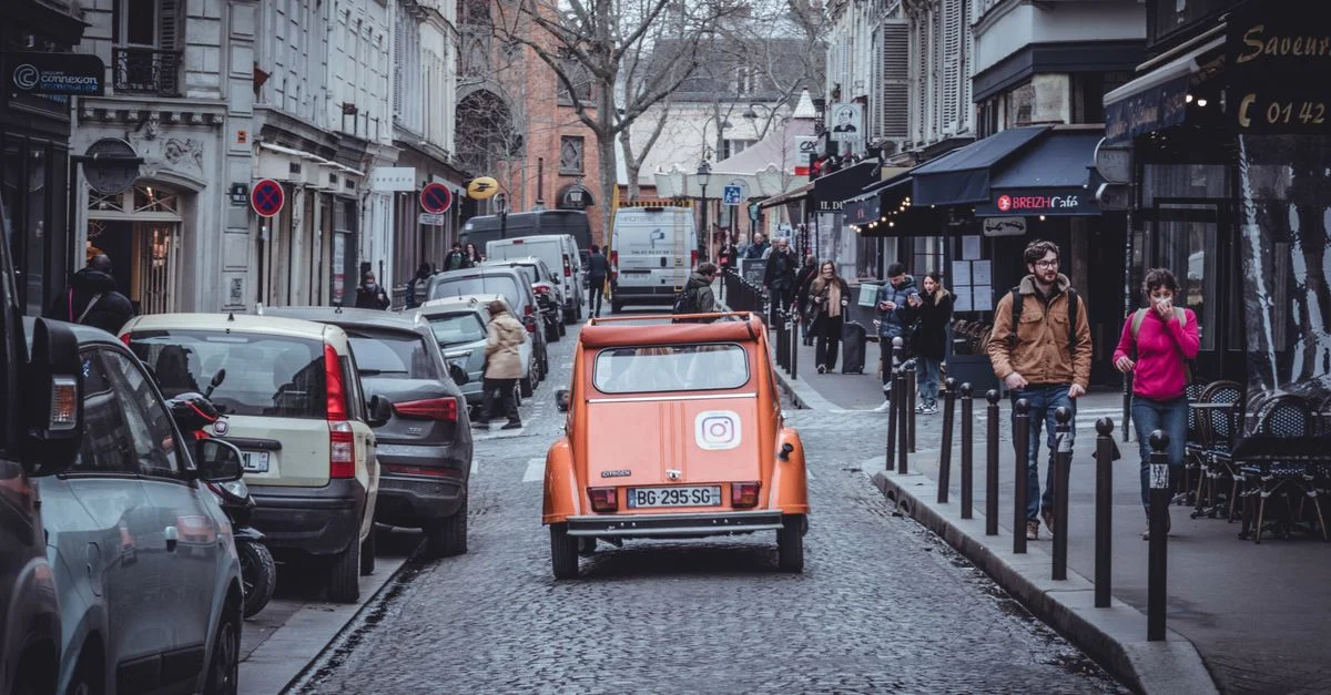 Une voiture vintage orange roulant sur une rue pavée parisienne bordée de cafés et de piétons.