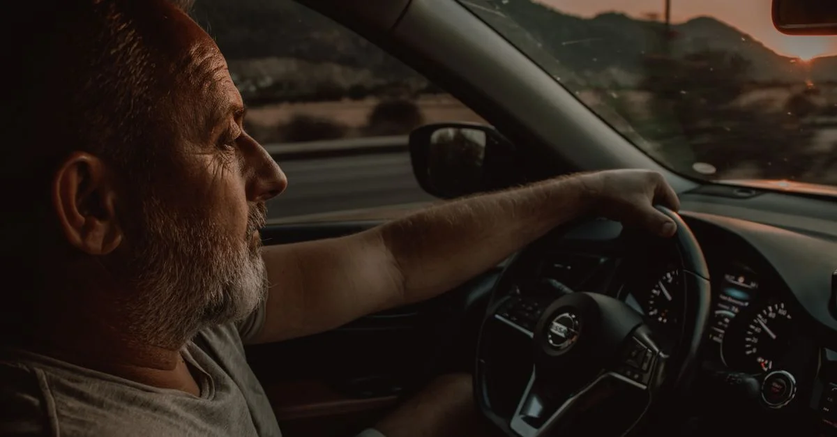 Un homme âgé conduisant une voiture Nissan pendant le coucher de soleil, illustrant le voyage et la contemplation.