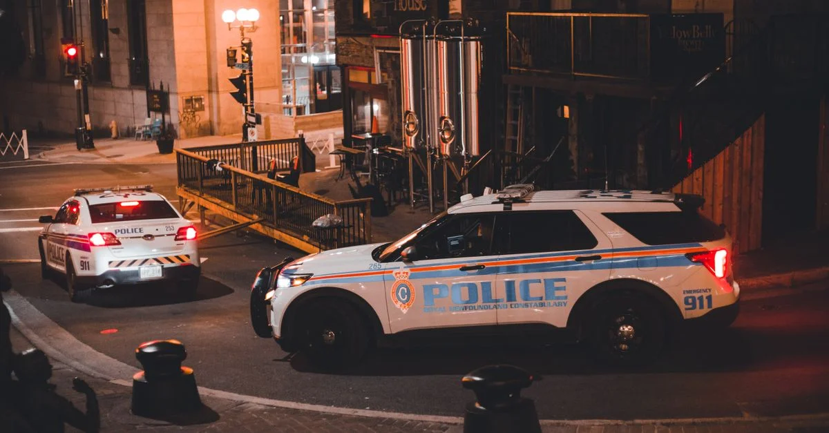Deux voitures de police patrouillant une rue de la ville la nuit avec un éclairage chaud créant une ambiance urbaine dramatique.