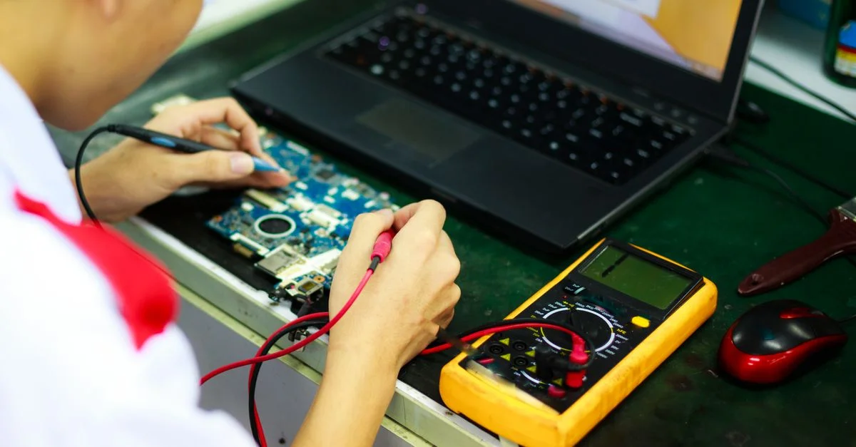 Technicien travaillant sur un ordinateur portable avec un multimètre dans un espace de travail professionnel.