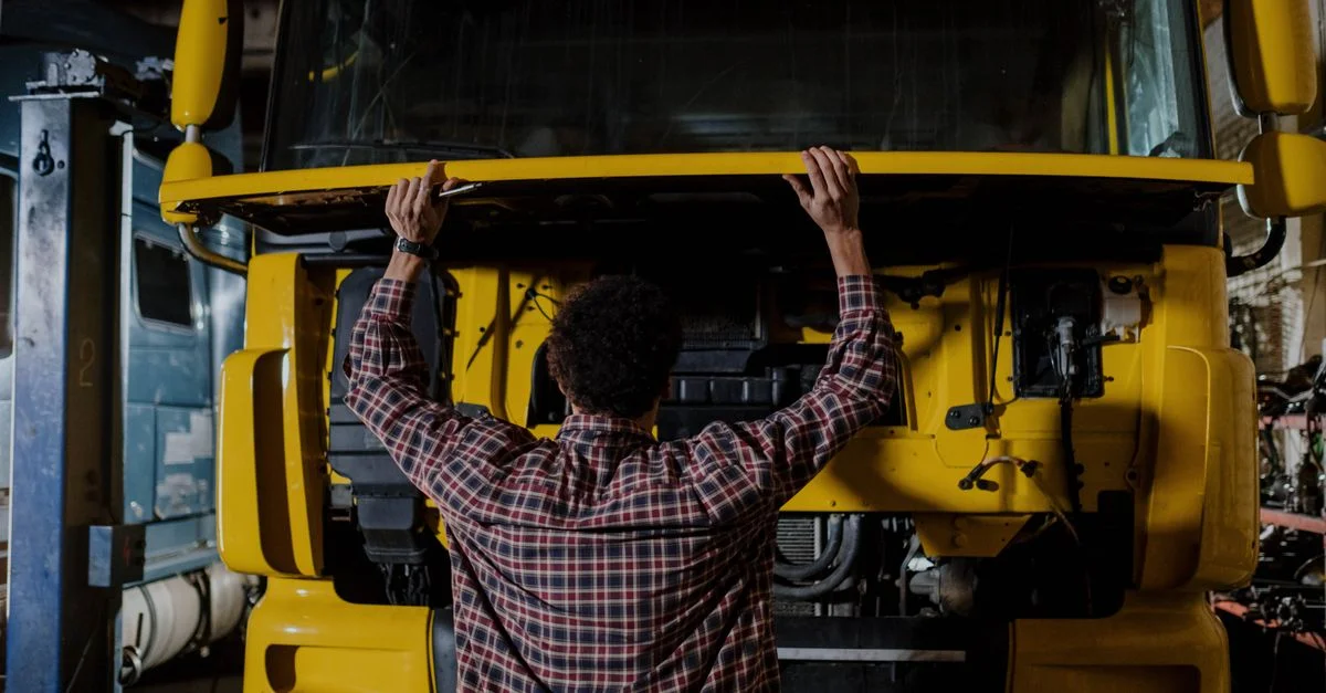 Vue arrière d'un mécanicien inspectant le moteur d'un camion jaune dans un garage.