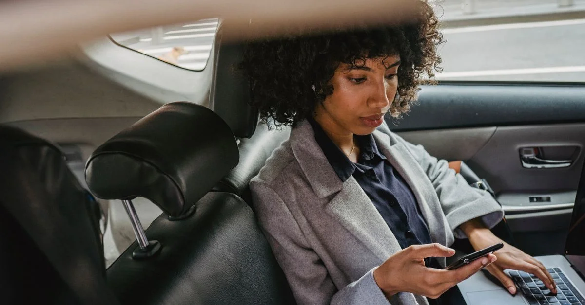 Femme multitâche avec un téléphone et un ordinateur portable sur le siège arrière de la voiture, équilibrant travail et voyage.