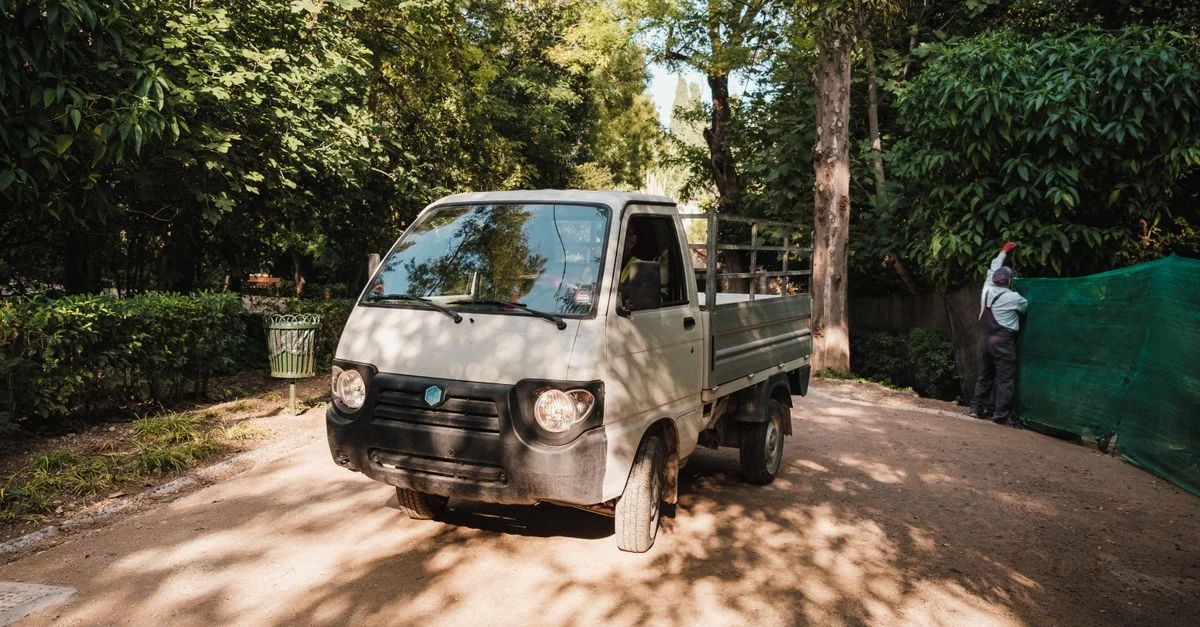 Petit camion blanc garé par une journée ensoleillée dans un parc verdoyant avec des arbres et un homme travaillant.