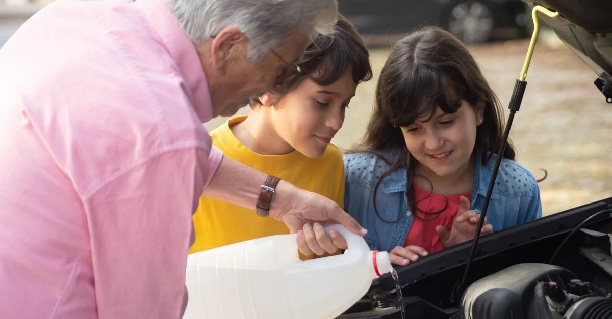 Un grand-père guide ses petits-enfants pour verser un liquide dans un moteur de voiture en extérieur.